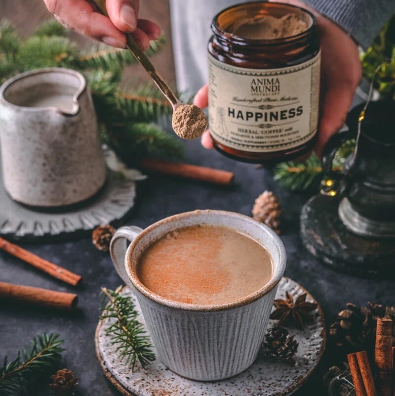 Im Hintergrund ein offens Anima Mundi Happiness Schraublas, davor ein Löffel mit Pulver gefüllt streut Pulver in eine gefüllte Kaffeetasse.