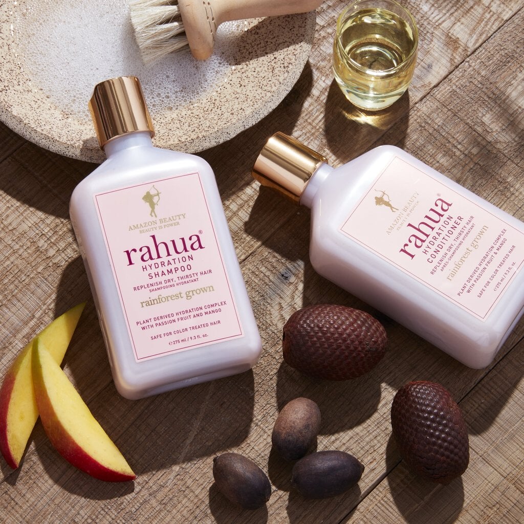 2 Rahua Flaschen Hydration Shampoo und Conditioner in Nahaufnahme liegen auf einem Tisch mit Rahuanuss, Obstspalten und Öl im Glas.