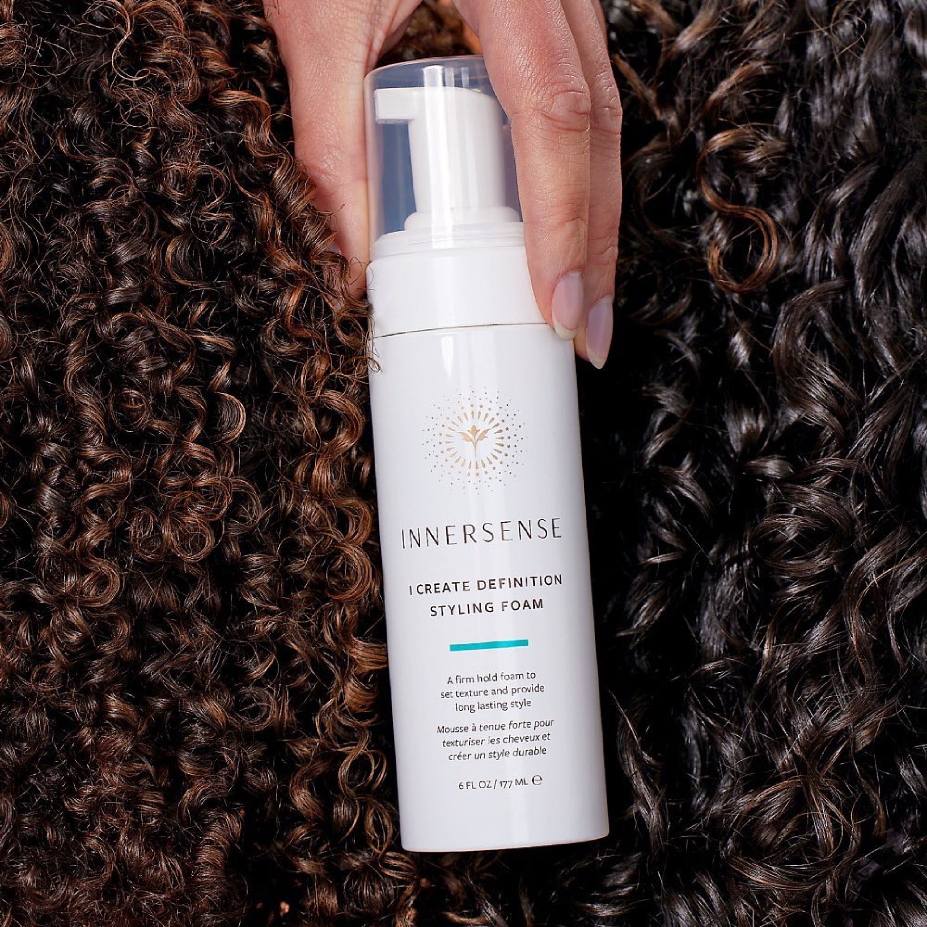 INNERSENSE I Create Definition Pumpflasche gehalten von einer Frauenhand vor stark gelockten Haaren.