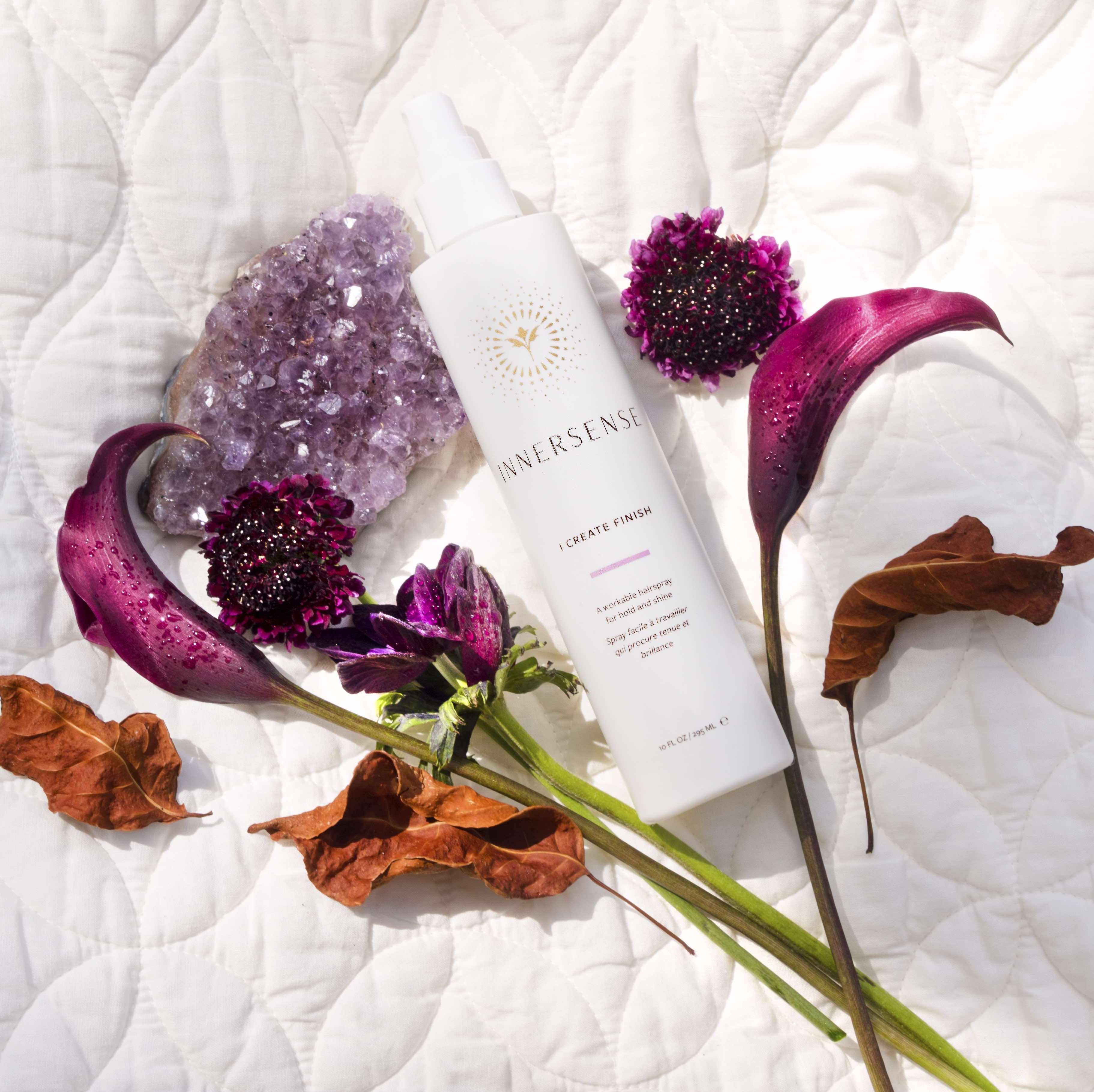 Auf einer weißen Decke liegt eine Innersense I Create Finish Sprühflasche zwischen pinkem Kristll, pinken Blüten und getrockneten Blättern