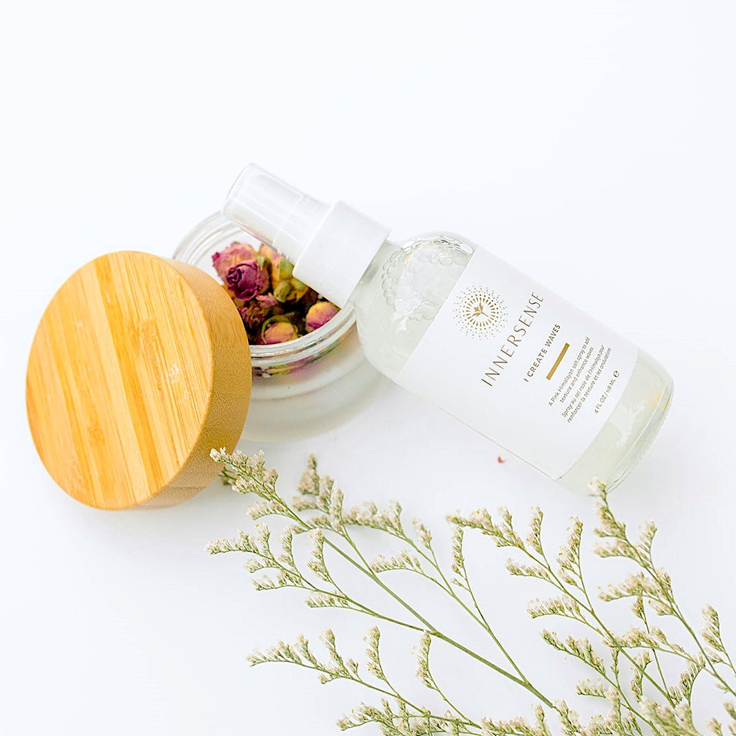 Innersense I Create Waves Sprühflasche liegt schräg auf einem Glas mit getrocknetem Rosenblüten auf weißem Untergrund.