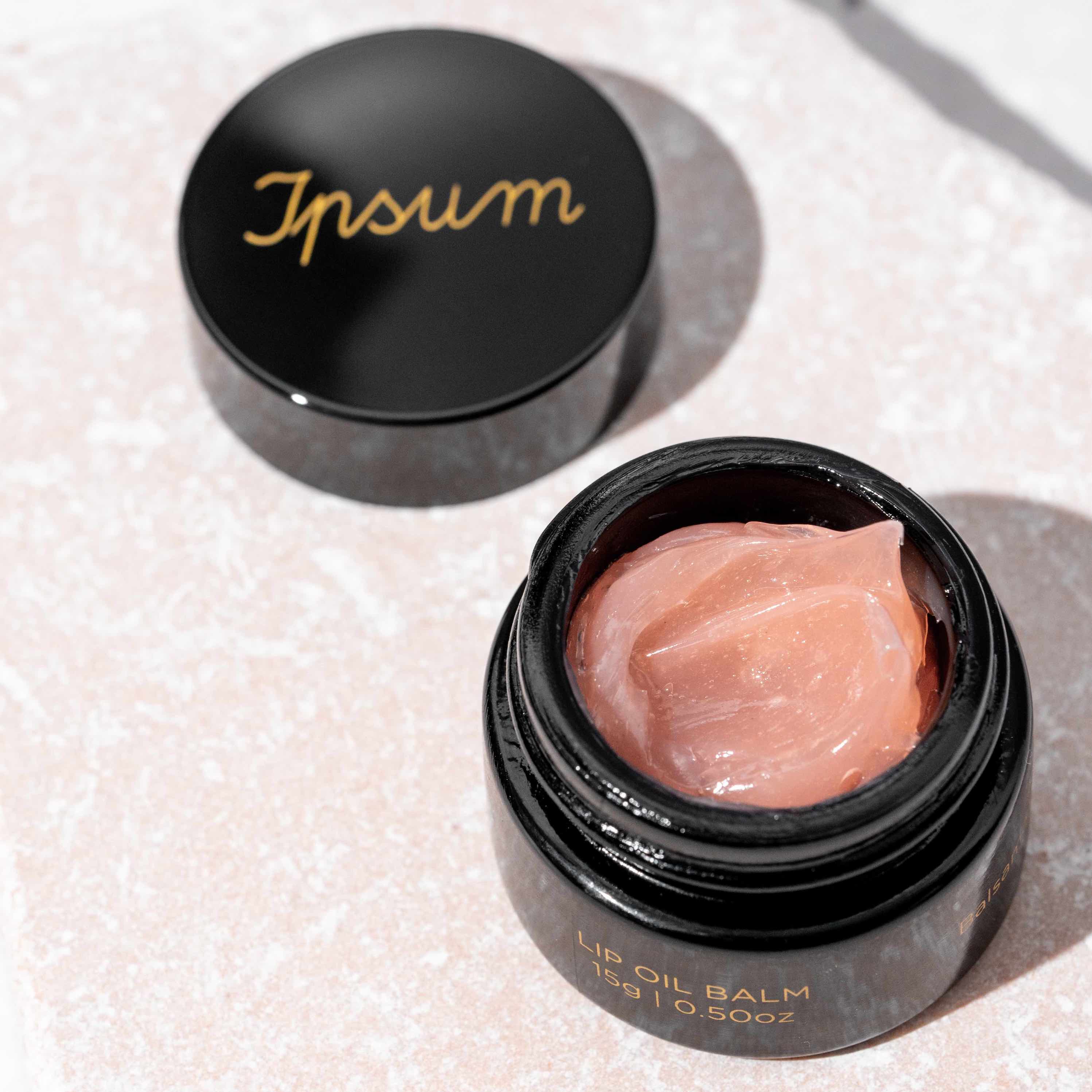 Schwarzer Ipsum Lip Oil Balm Tiegel steht offen, daneben liegt der Deckel, Balm im zarten Rosa und geschmeidiger Textur gut zu erkennen.