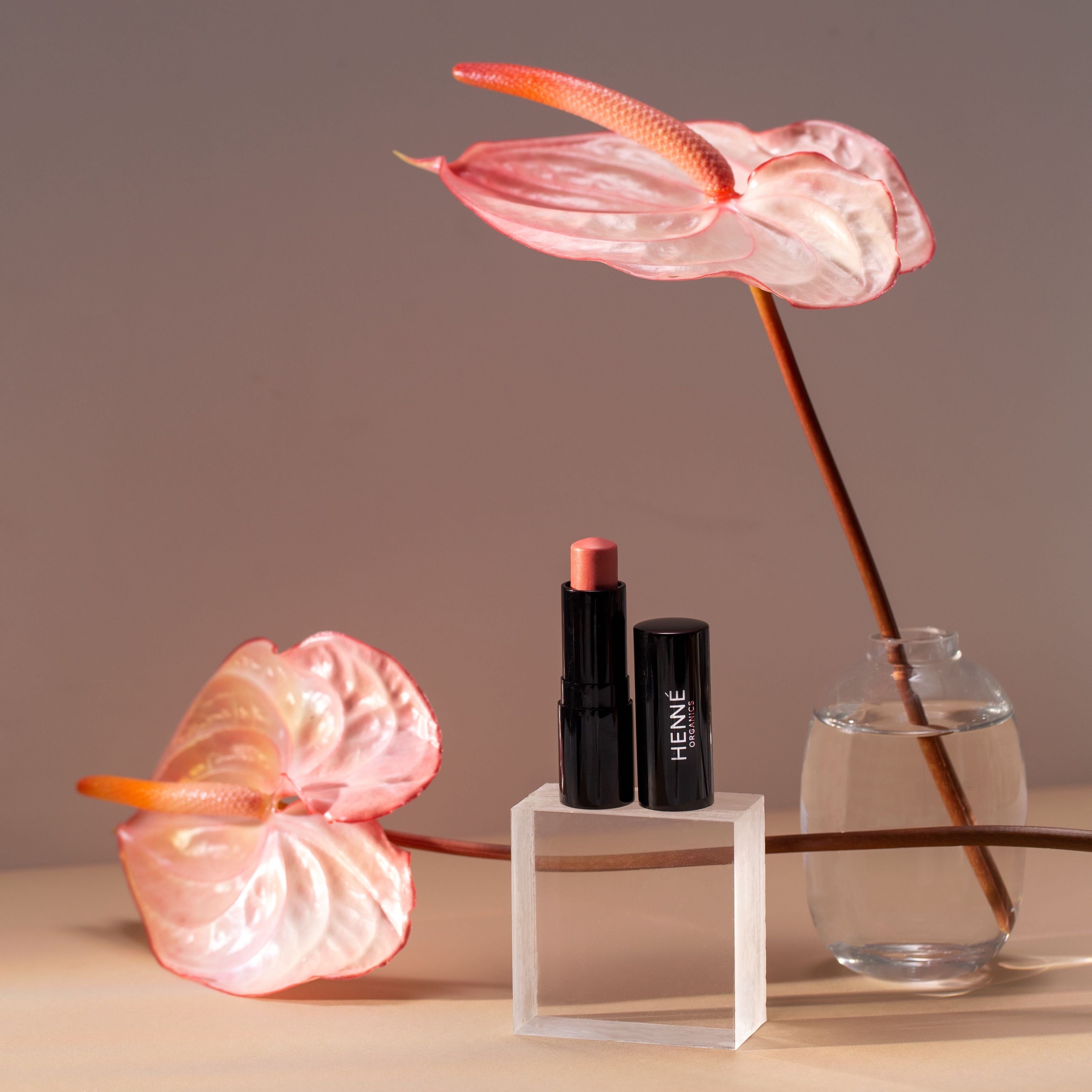 Schwarzer Lippenstift mit Farb- und Texturbeispiel der Farbe Azalea von Henné Organics steht zwischen 2 Blumen.