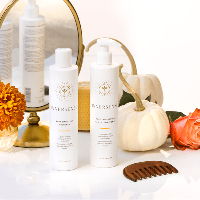 2 Flaschen Innersense Pure Harmonny Hairbath und Pure Inspiration Daily Conditioner stehen zwischen einem kleinen Standspiegel, Blumenvase mit orangefarbener Blüte, Holzkamm, weißem Kürbis und Rosenblüte vor weißem Hintergrund.