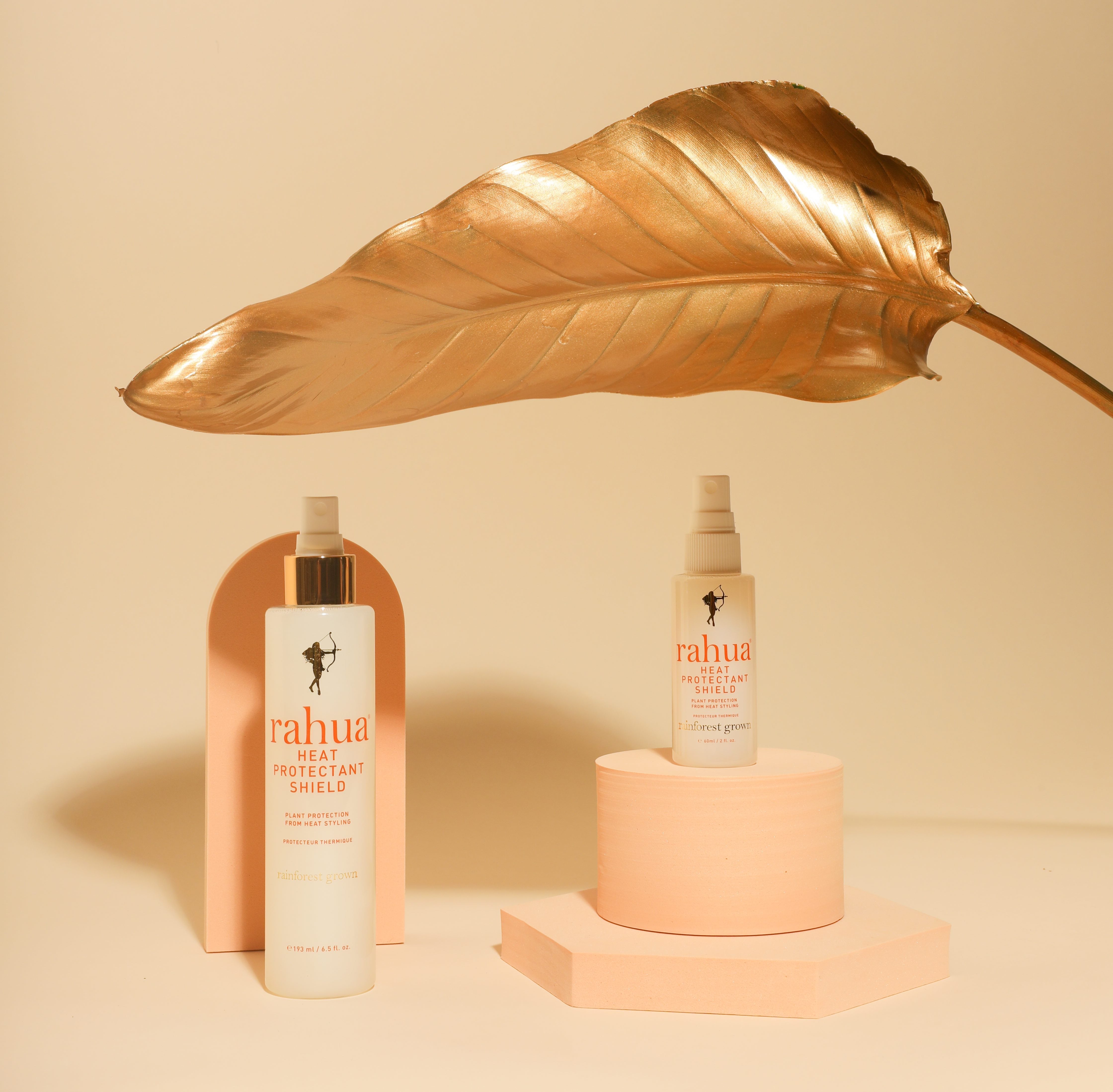 2 Flaschen Rahua Heat Protection Shield in Original- und Travel-size-größe unter einem goldenem Palmenblatt.