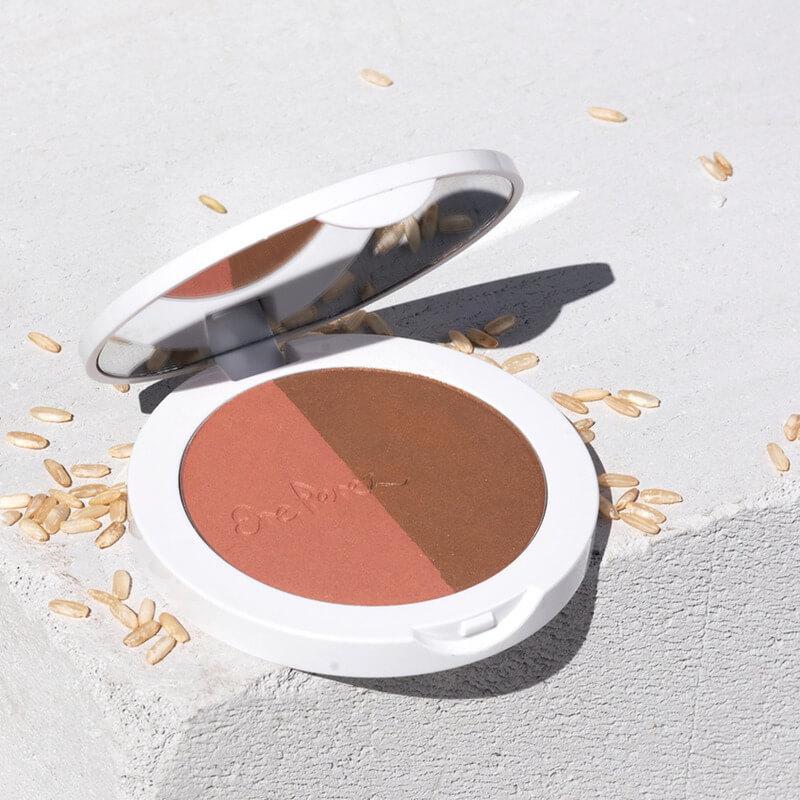 Offene Dose des Rice Powder Blush & Bronzer BROOKLYN auf hellem Untergrund mit Reiskörnern.