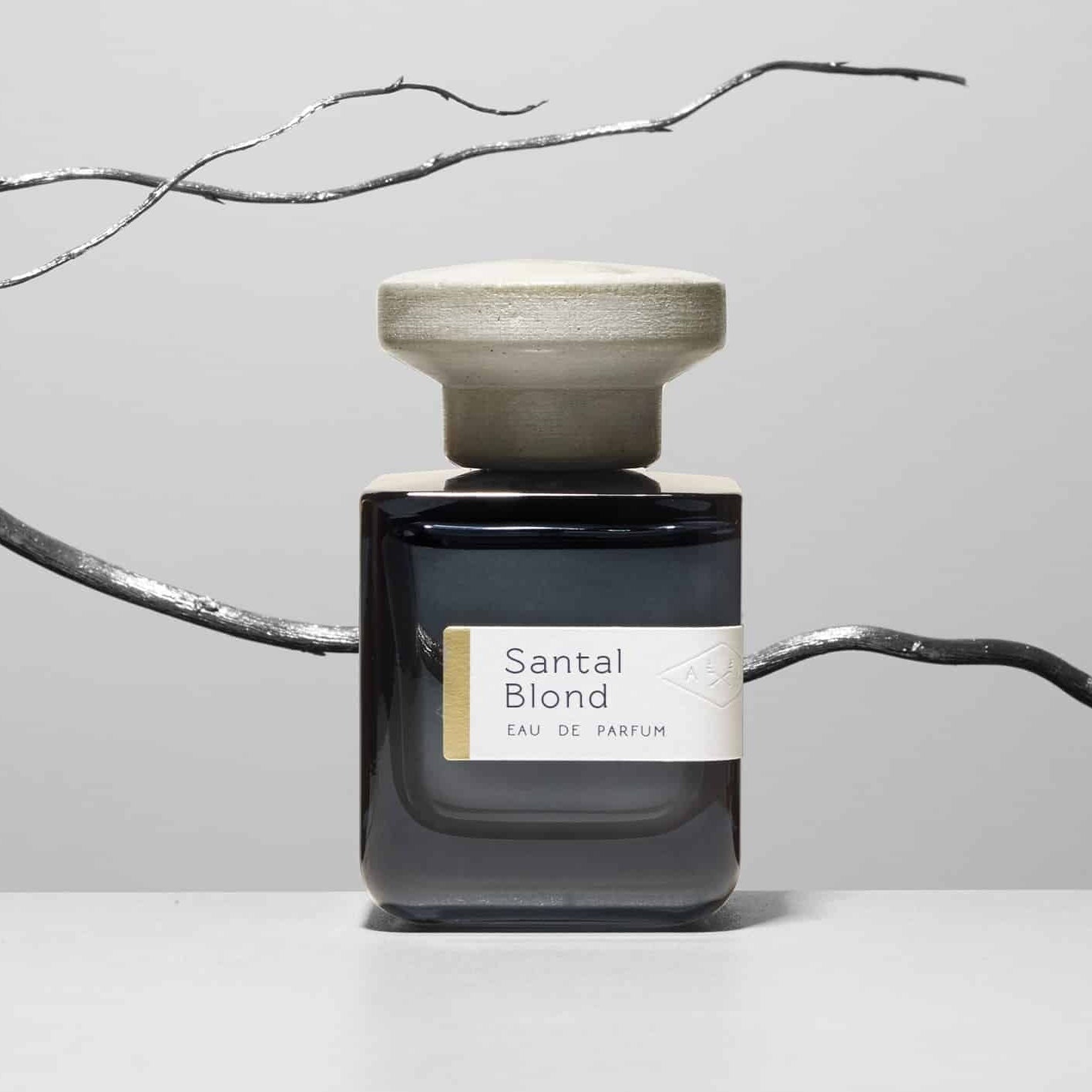 Dunkelblaue Glasflasche mit grauem Deckel "Santal Blond" von Atelier Materi mit einem silbernen Ast im Hintergrund.