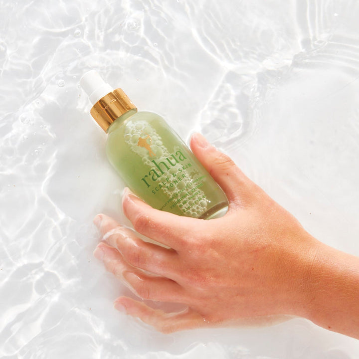 Frauenhand hält eine liegende Rahua Scalp & Skin Toner Flasche im Wasser vor hellem Hintergrund.