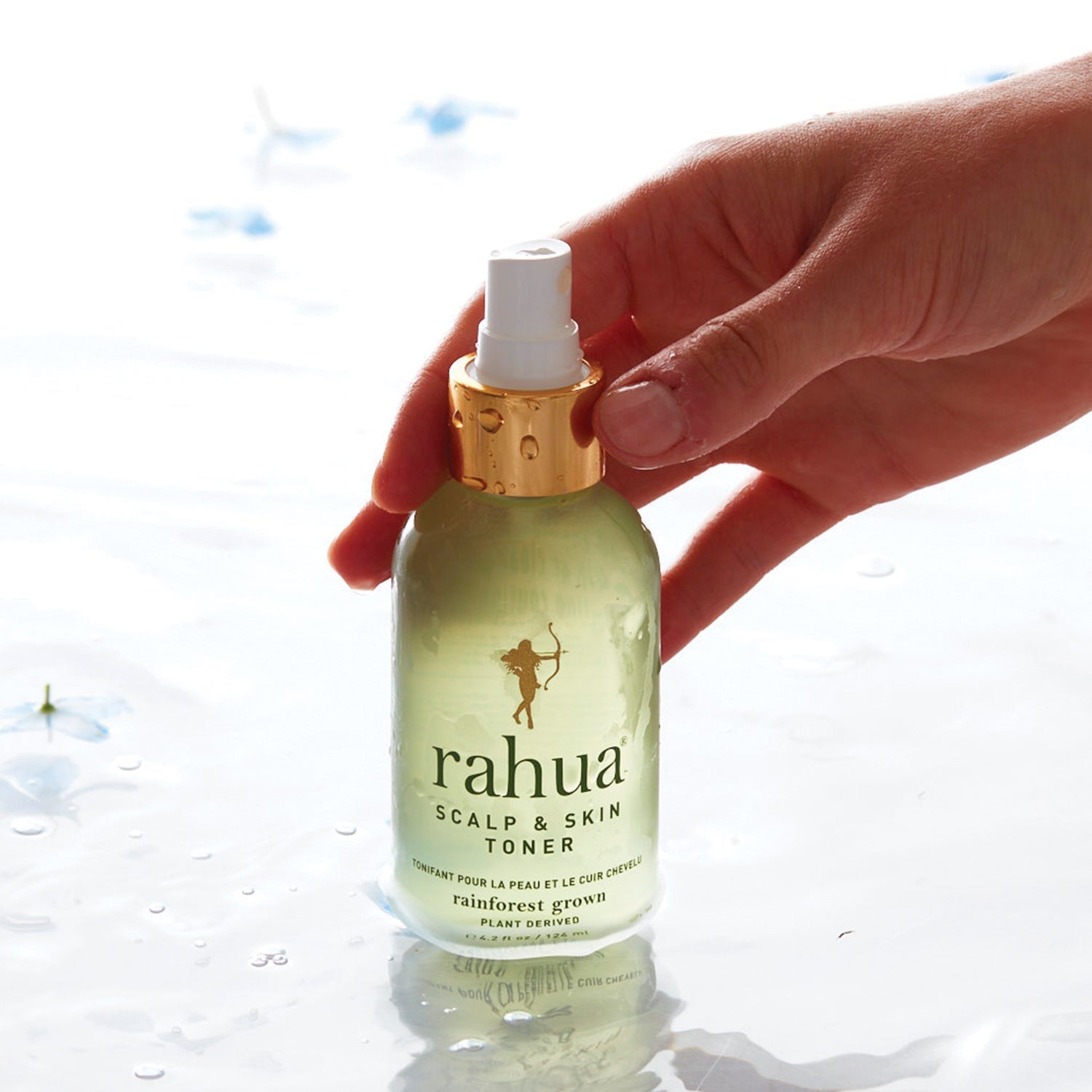 Frauenhand hält eine stehende Rahua Scalp & Skin Toner Flasche im Wasser vor hellem Hintergrund.