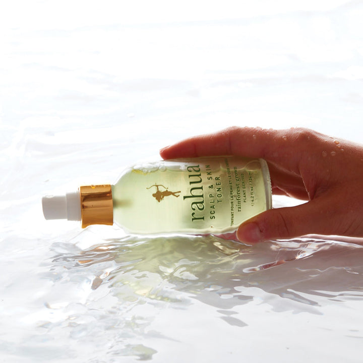 Frauenhand hält eine umgekippte Rahua Scalp & Skin Toner Flasche im Wasser vor hellem Hintergrund.