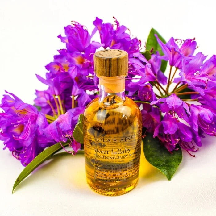 Glasflasche Lola´s Apothecary mit orngefarbenem Inhalt, dahinter lilafarbene Blumen.