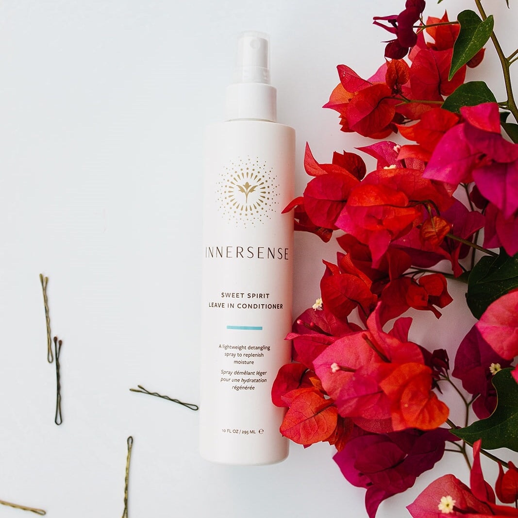 Innersense Sweet Spirit Leave In Conditioner Sprühflasche liegt mittig auf weißem Hintergrund neben verstreuten Haarnadeln und Blumendekoration aus der Vogelperspektive.