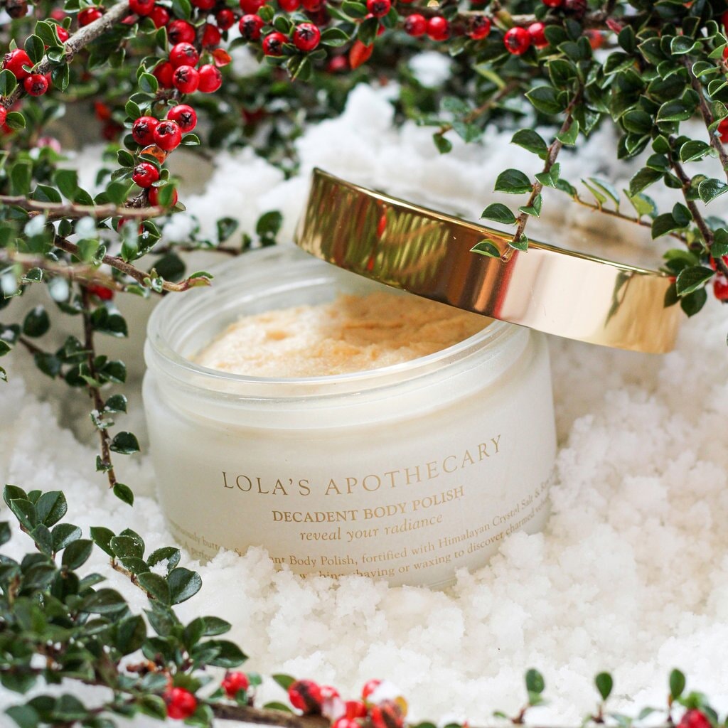 Lola's Apothecary Glastiegel Body Polish mit goldenem Deckel, der offen gegen das Glas lehnt, steht im Schnee umringt von roten Beeren und Zweigen.
