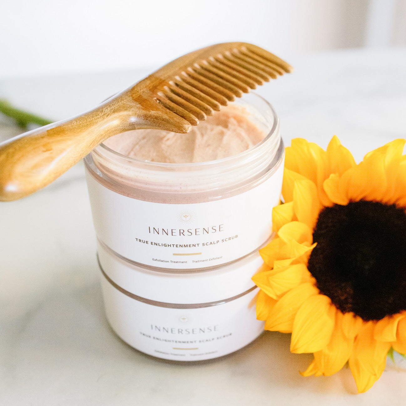 2 Innersense True Enlightenment Scalp Scrub Tiegel stehen aufeinenader, der obere offen und drauf liegt ein Holzkamm neben einer Sonnenblume.