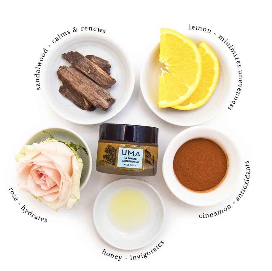 UMA Ultimate Brightening Face Mask Schraubglas mit schwarzem Deckel liegt in der Mitte umringt von den Zutatet Sandelholz, Zitronenscheien, Rosenblüte, Schälchen Honig und einem Schälchen Zimtpulver.