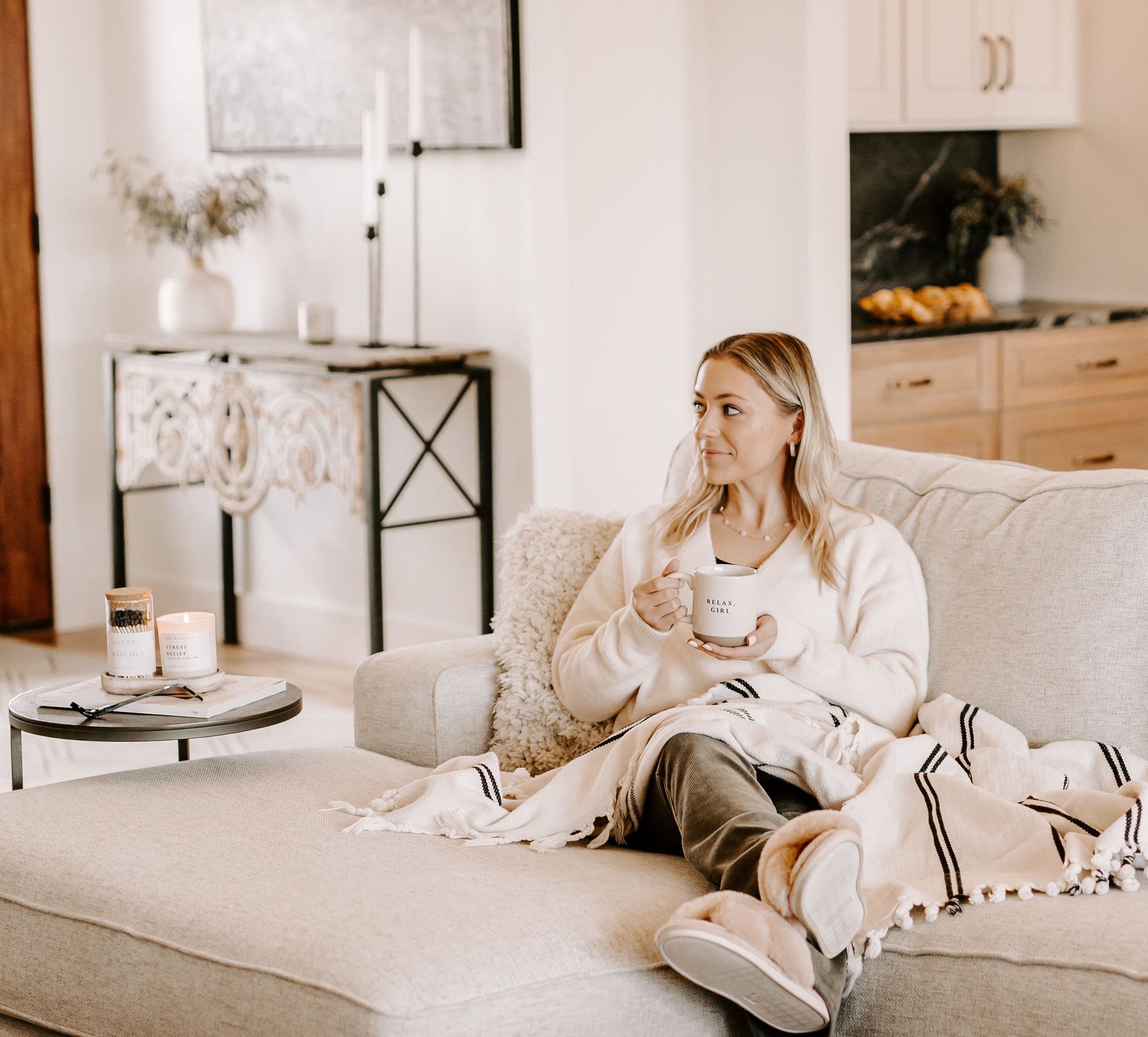 Woman sitting on a couch in a cozy atmosphere next to soy wax candles and matches from Sweet Water Decor, holding a cup from Sweet Water Decor in her hands.
