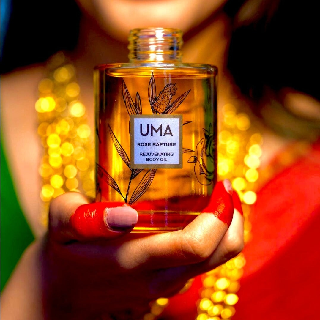 Woman blurred in the background with colorful clothes and many gold chains holding the UMA Rose Rapture Rejuvenationg Body Oil bottle in the foreground.