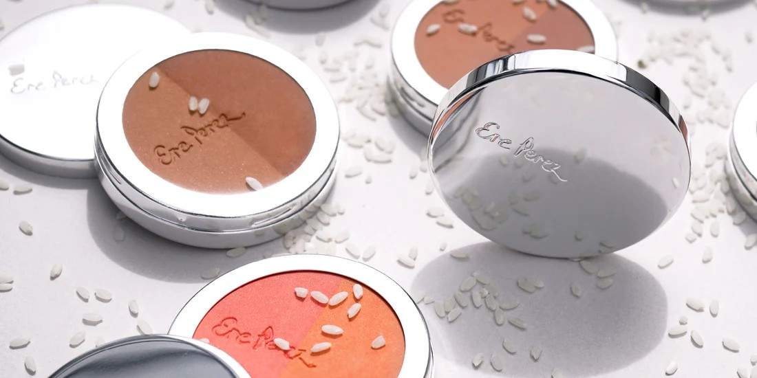 Verschiedene Bronzer und Blushes von Ere Perez liegen geöffnet auf weißem Untergrund mit verstreuten Reiskörnen.