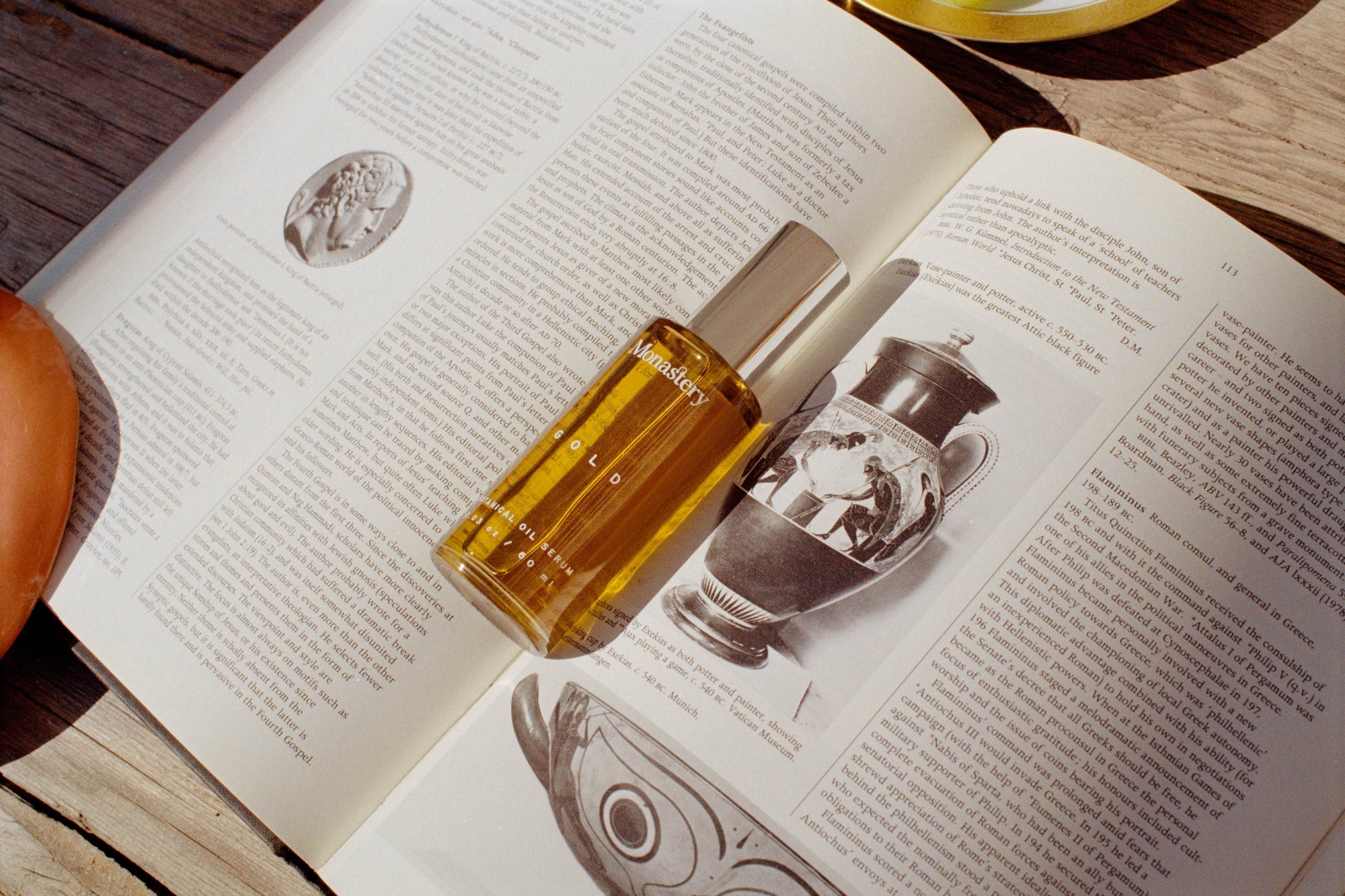 Monastery Gold Serum Flasche liegt auf aufgeschlagenem Buch auf einem Holztisch.
