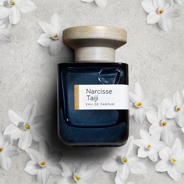 Parfumflasche Narcisse Taiji liegend auf weißem Sand mit Blüten von Narcissen