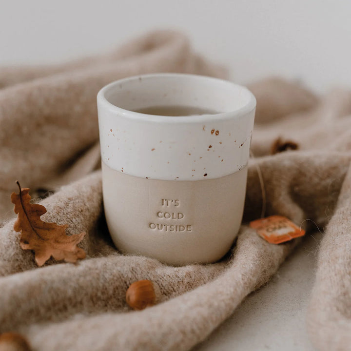 Kaffeebecher mit Prägung "it's cold outside" steht auf einer hellbraunen Decke umgeben von einem Eichenblatt und einer Eichel.