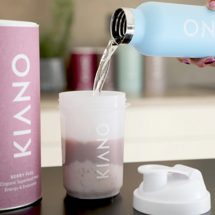 Wasser wird in einen Milchshakebereiter mit Kiano Berry Fuel Pulver gegossen, daneben steht eine Dose Kiano Berry Fuel auf einer Küchenarbeitsplatte