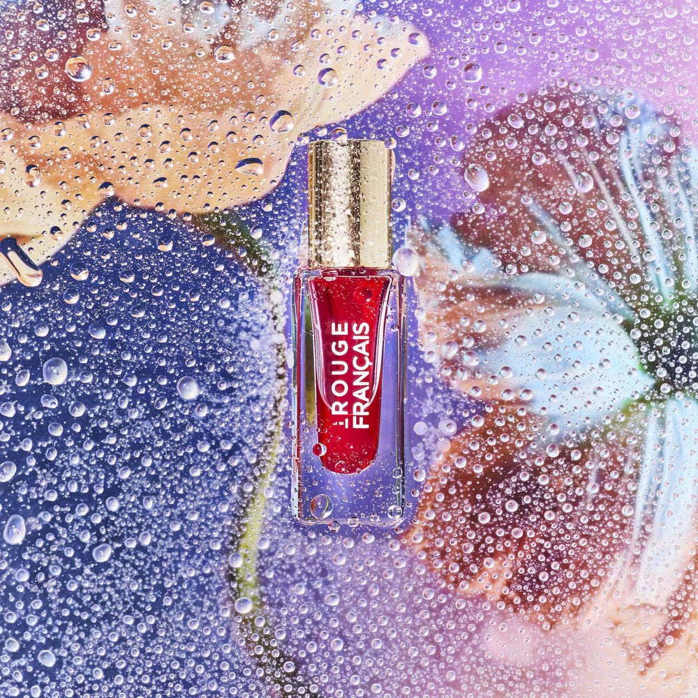 Lippenöl in einer Glasflasche mit goldenem Deckel von Le Rouge Francais, verschwommen im Hintergrund Blütenblätter und viele Wassertropfen.