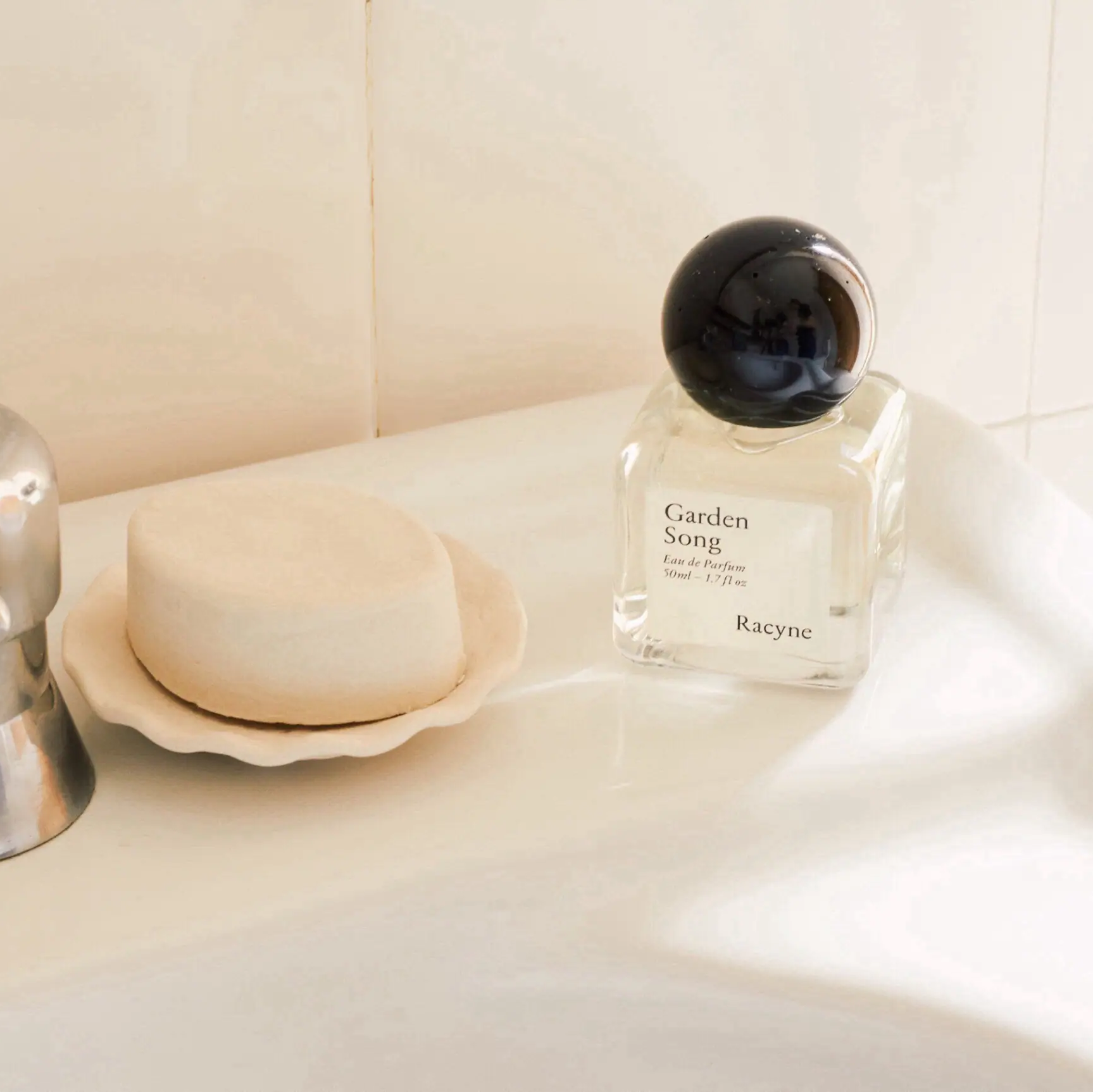 Garden Song Eau de Parfum von Racyne auf dem Rand eines Waschbeckens neben einer Schale mit Seife.