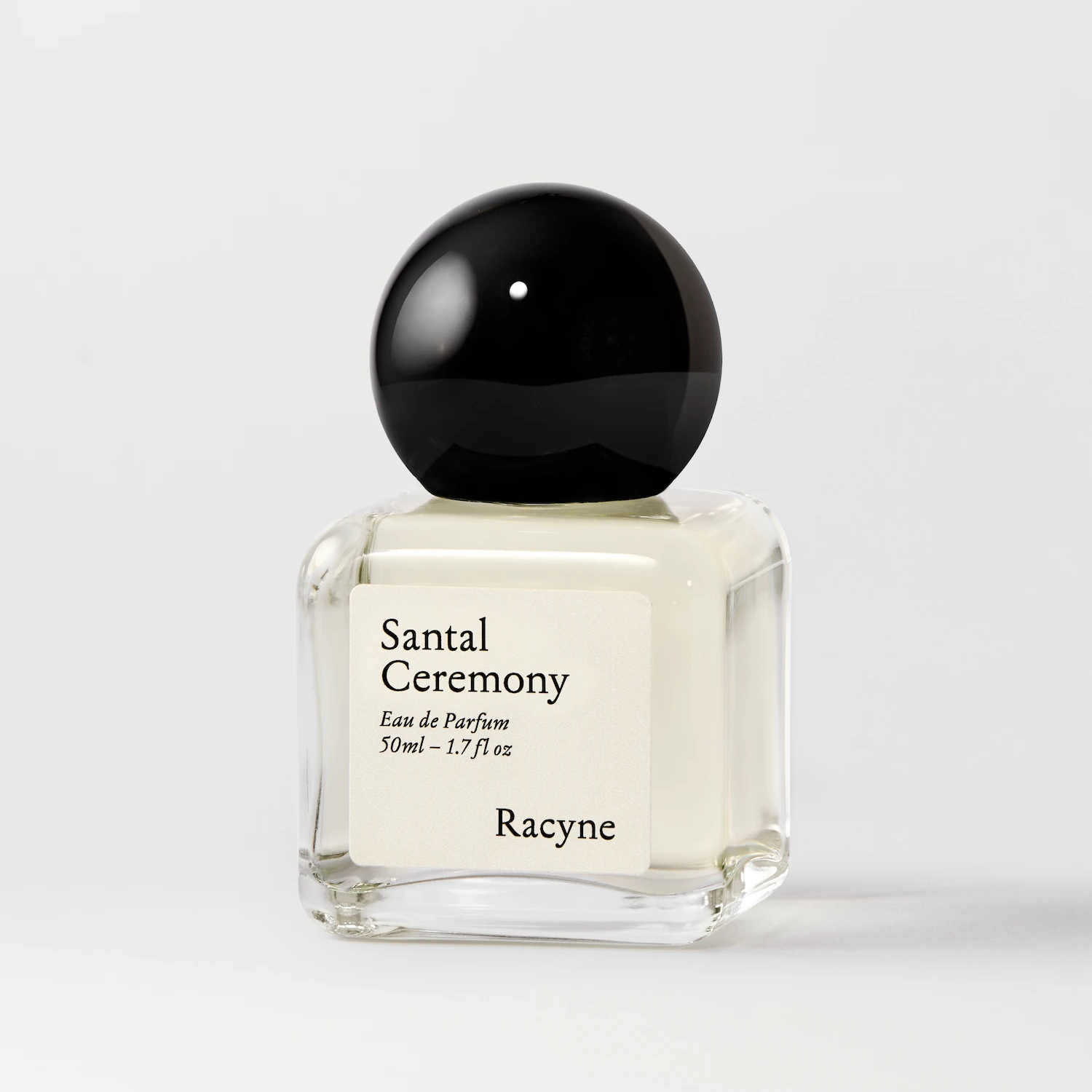 Santal Ceremony Eau de Parfum von Racyne – transparenter Parfumflakon mit cremefarbenem Etikett und schwarzem Kugelverschluss