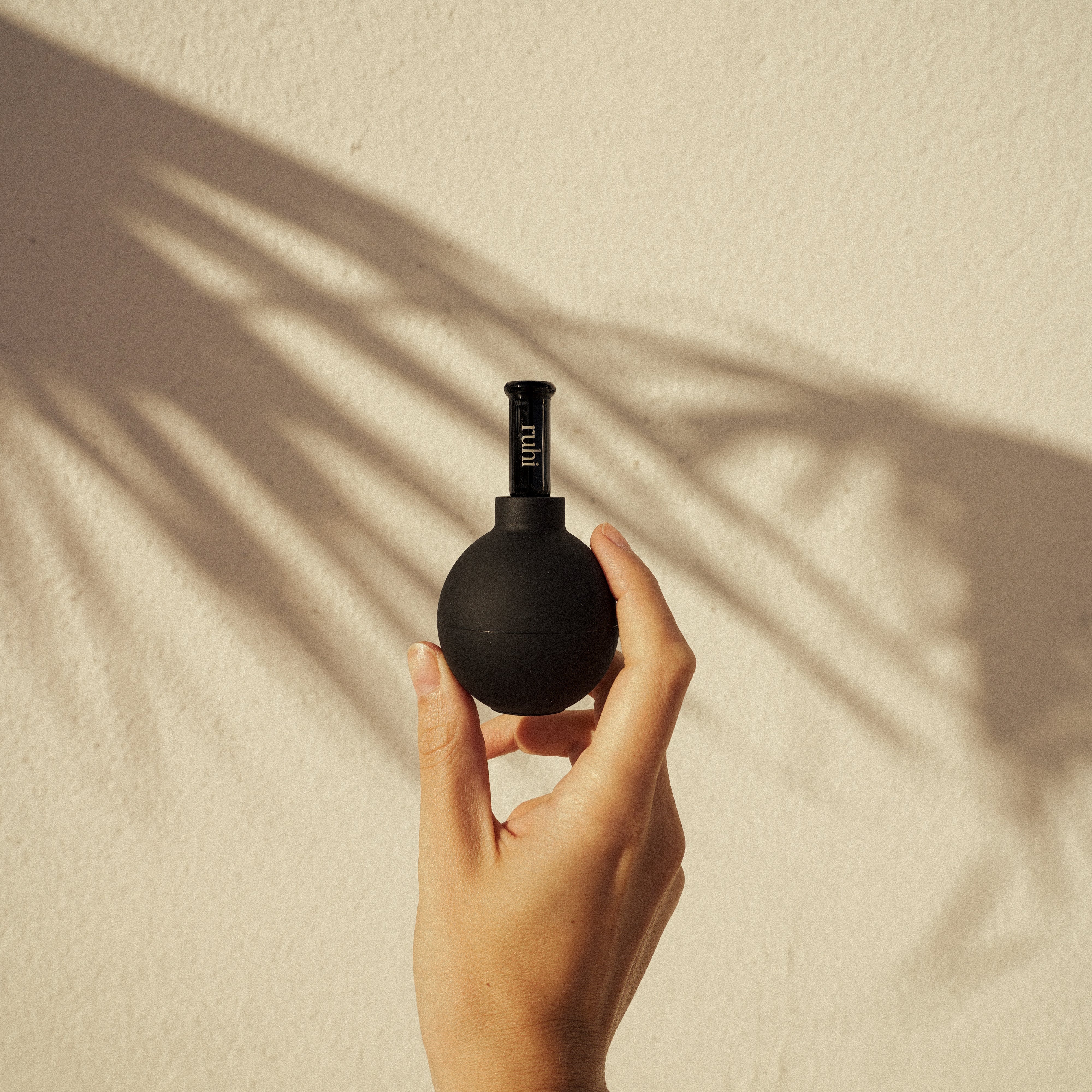  Ruhi The Facial Cups in einer Hand vor. Beigem Hintergrund mit Palmenschatten