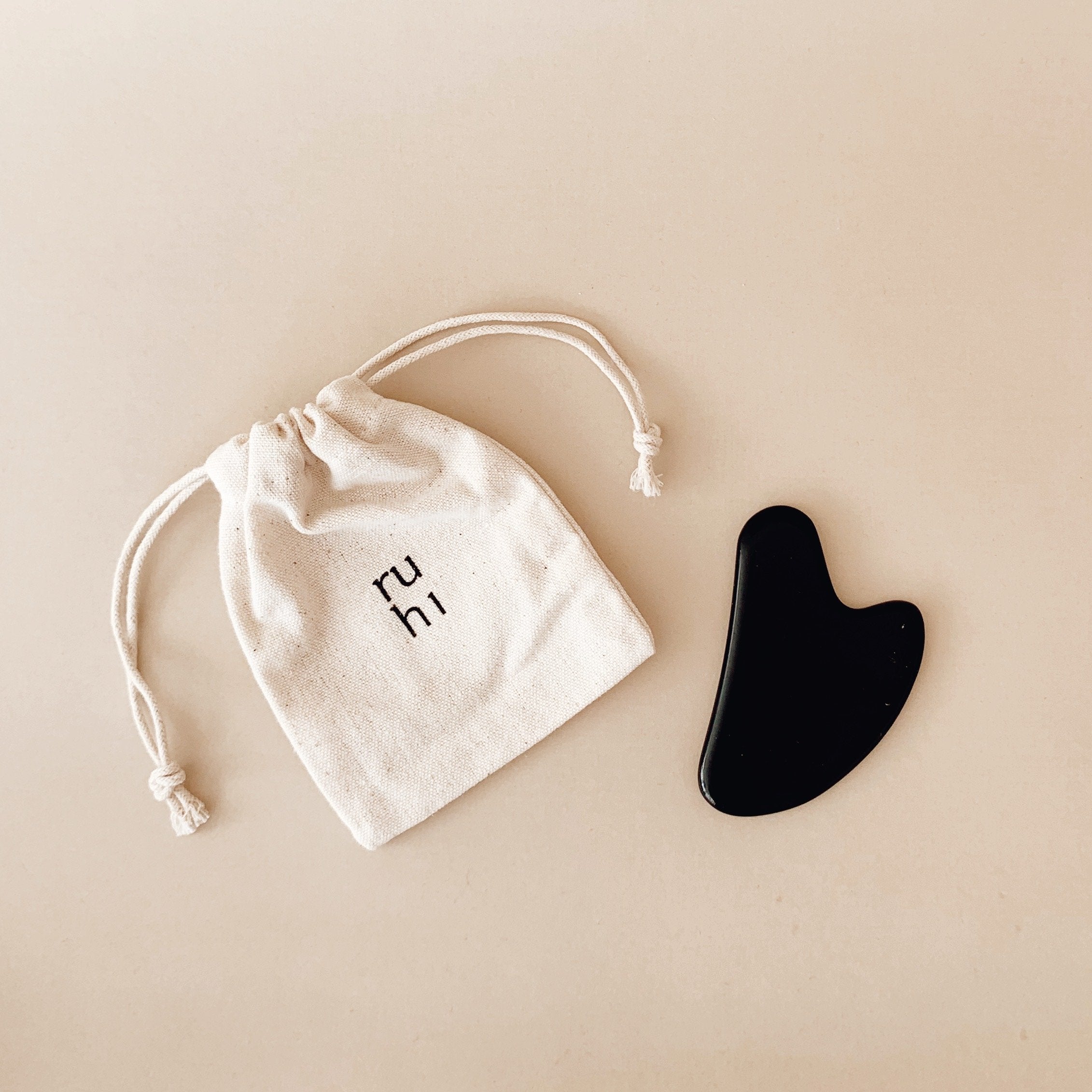 Ruhi The Obsidian Gua Sha mit Canvassäckchen vor beigem Hintergrund