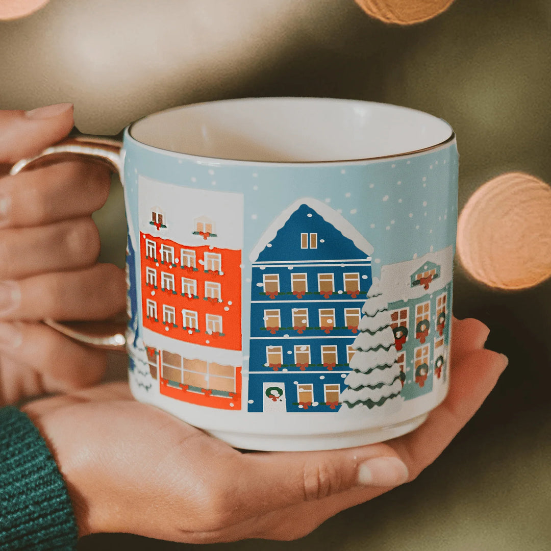 Weihnachtstasse "Christmas Village" von Sweet Water Decor wird von zwei Händen gehalten.