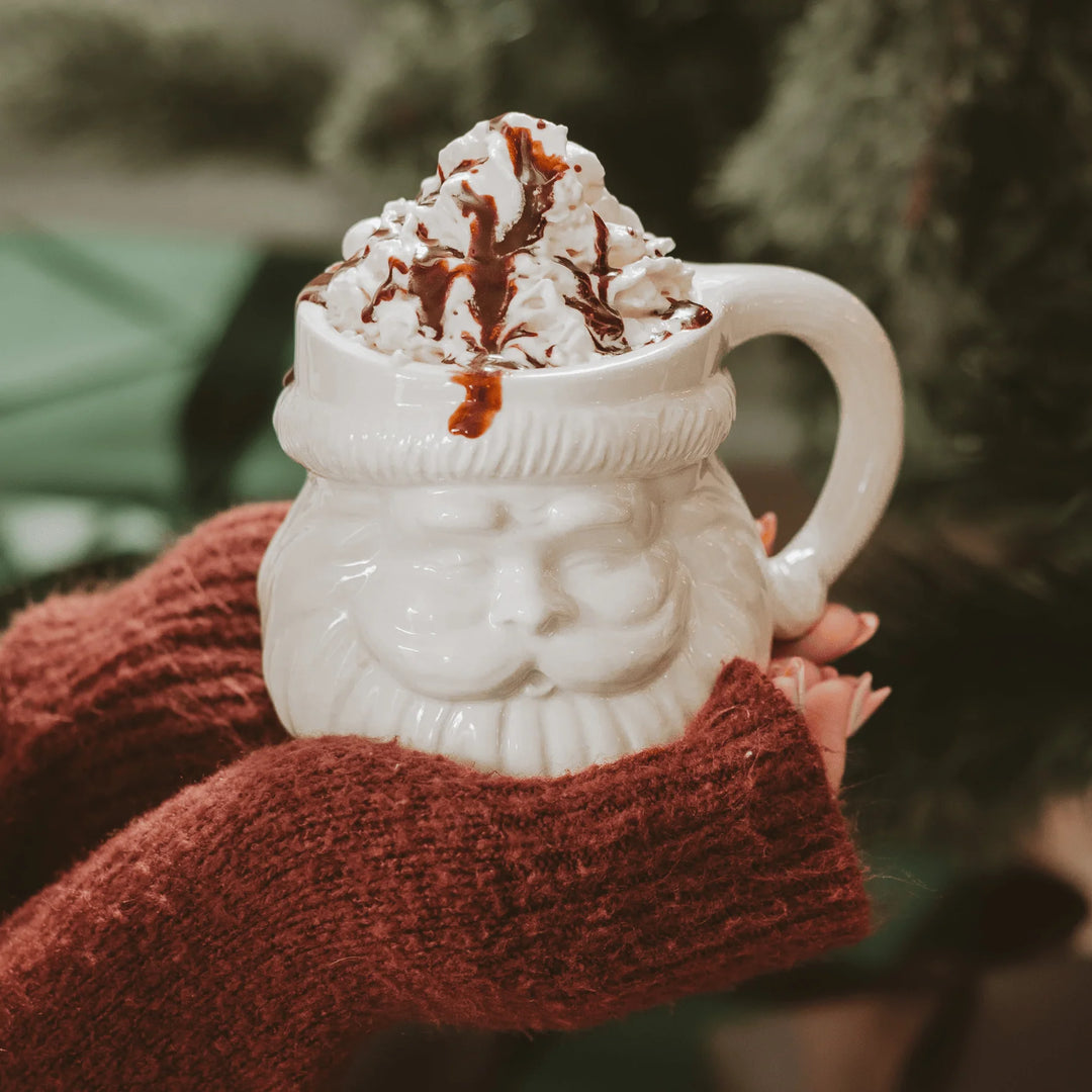 Cremefarbene Tasse mit Weihnachtsmanngesicht-Design von Sweet Water Decor, mit sahnigem Getränk, gehalten von zwei Händen. North Glow