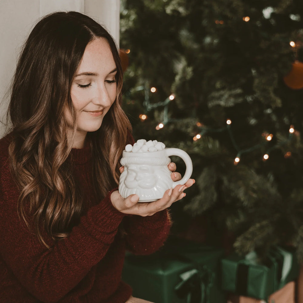 Cremefarbene Tasse mit Weihnachtsmanngesicht-Design von Sweet Water Decor wird von einer Frau mit langen Haaren in den Händen gehalten.