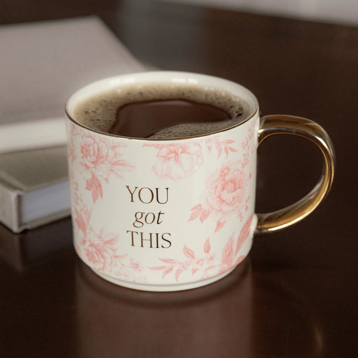 Tasse mit rosa Muster, goldener Aufschrift "YOU got THIS" und goldenem Henkel, gefüllt mit Tee, stehen auf einem braunen Tisch.