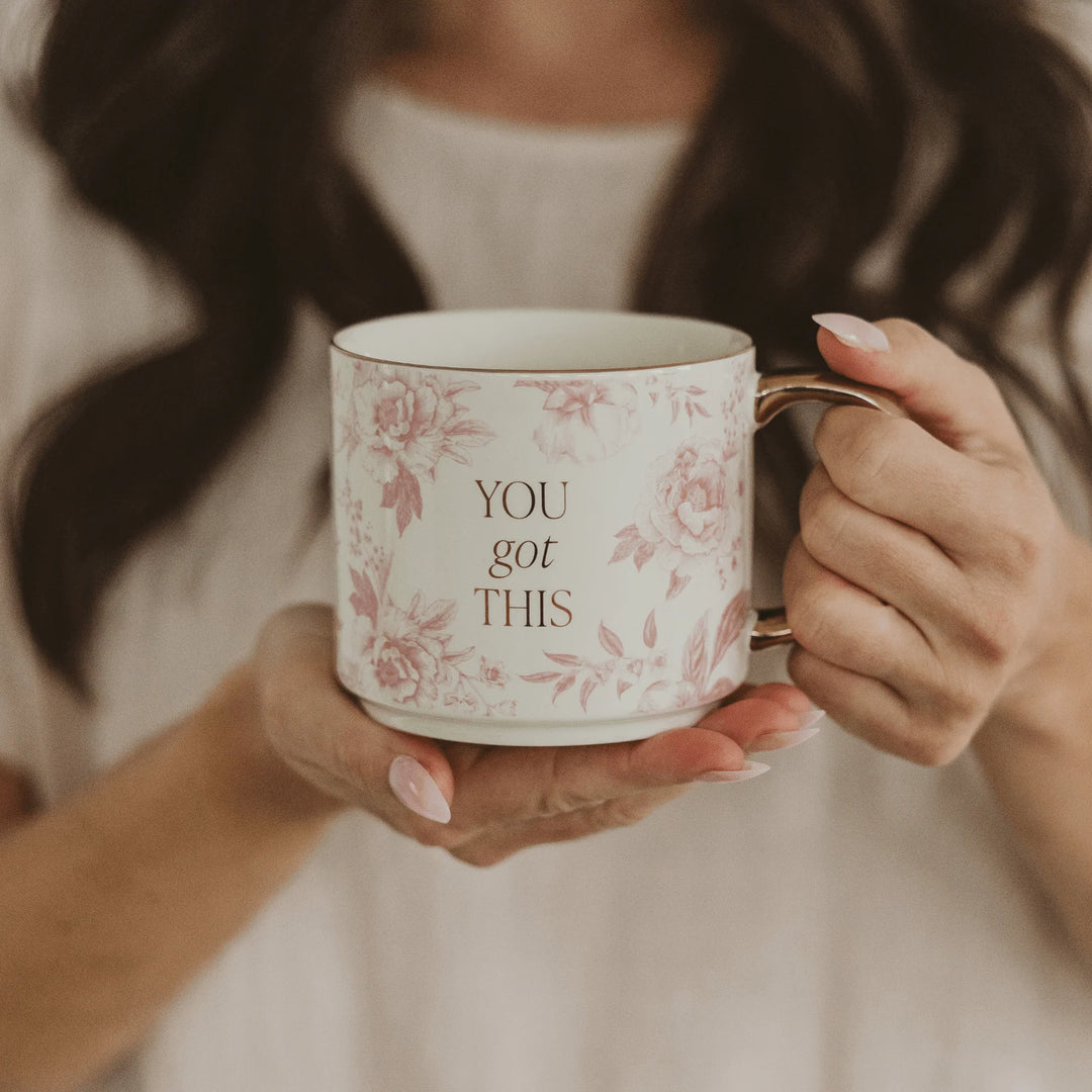 Tasse mit rosa Muster, goldener Aufschrift "YOU got THIS" und goldenem Henkel, wird von zwei Händen gehalten.