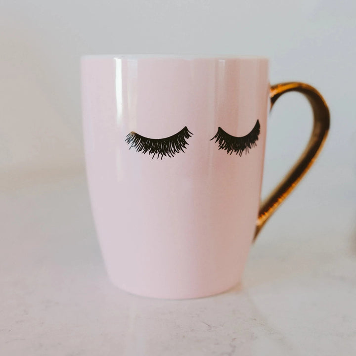 Rosafarbene Kaffeetasse "Pink Eyelashes" mit schwarzen Wimpern und goldenem Henkel vor hellem Hintergrund