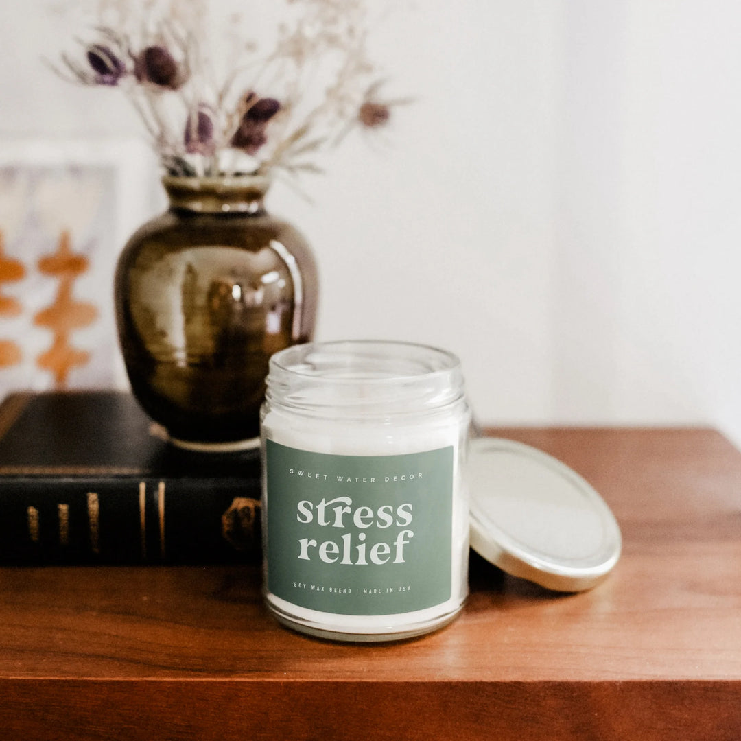 Geöffnetes Kerzenglas "stress relief" von Sweet Water Decor steht auf einem braunen Holztisch, daneben getrocknete Blumen in einer Vase und ein Buch. North Glow