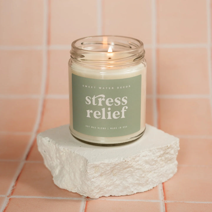 Kerzenglas "stress relief" von Sweet Water Decor steht auf einem Stein, der auf einem rosafarbenem Fliesenboden steht.