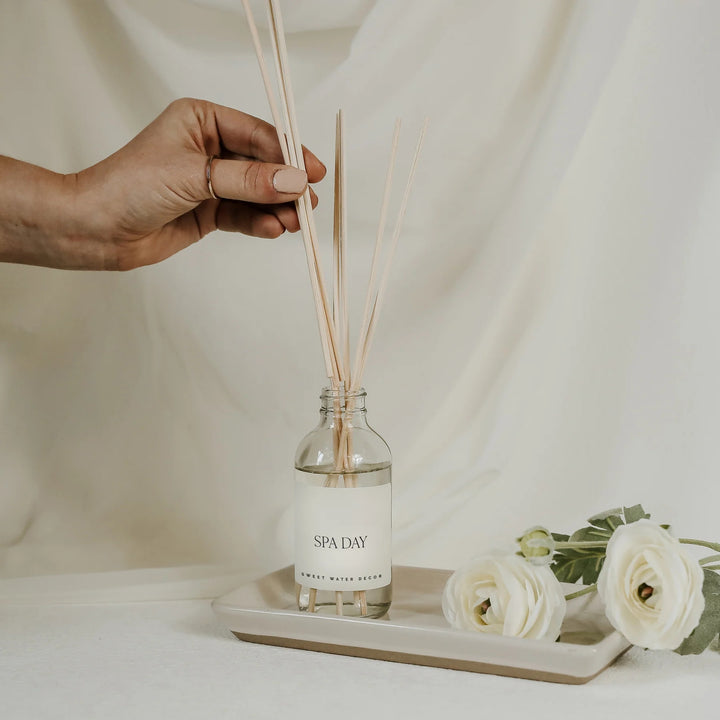 Hand hält Schilfrohrstäbchen in eine Flasche Sweet Water Decor Diffuser Spa Day, die auf einem Tablett steht mit weißen Blumen vor hellem Hintergrund.