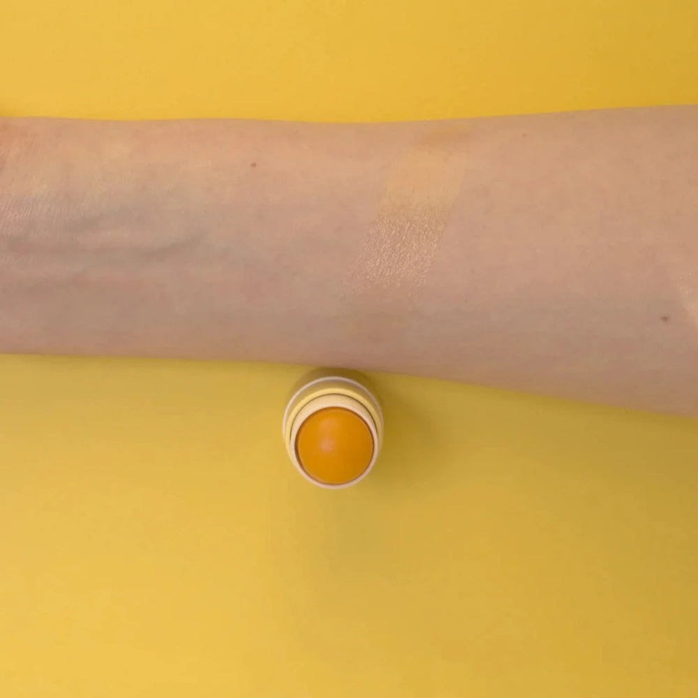 Ein Arm und ein geöffneter "The Balm" von PURE U. Cosmetics vor gelbem Hintergund.