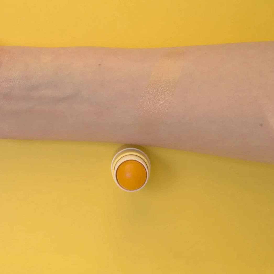 Ein Arm und ein geöffneter "The Balm" von PURE U. Cosmetics vor gelbem Hintergund. North Glow