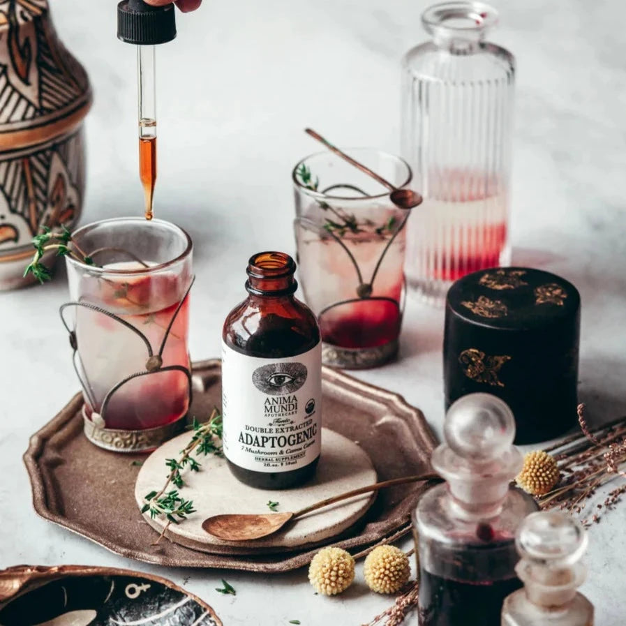 Anima Mundi Glasflasche Adaptogenic steht offen zwischen Gläsern, Löffeln, Flaschen und Blütendekoration.