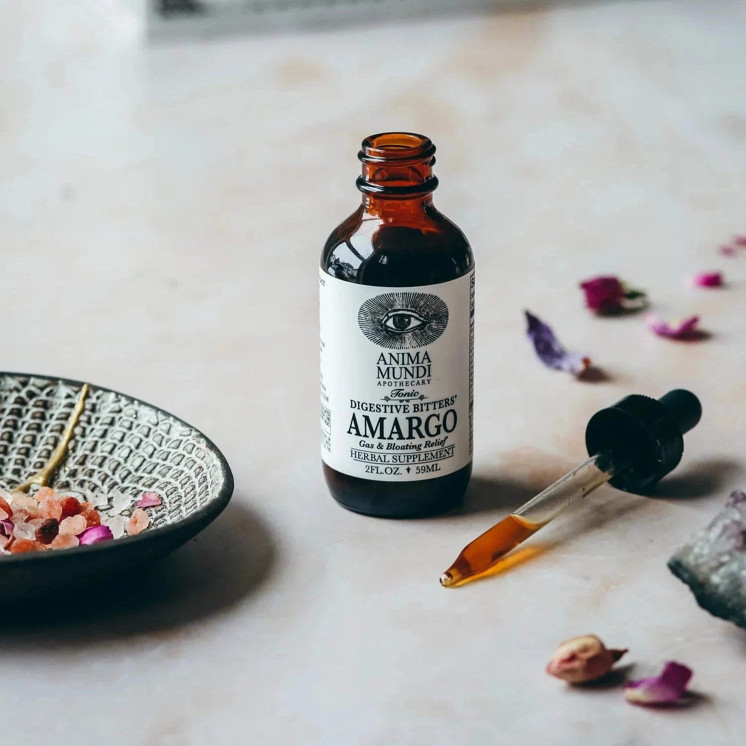 Anima Mundi Glasflasche Amargo mit daneben liegender Pipette vor hellem Hintergrund zwischen getrockneten Blütenblättern.