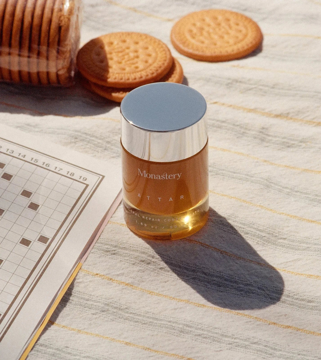 Monastery Gold Balm Konzentrat Tiegel mit 50ml Inhalt auf Badetuch mit Rätselheft und Keksen vermittelt Urlaubsfeeling. North Glow