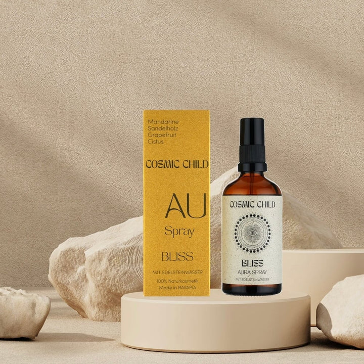 Verpackung und braune Sprühflasche von Cosmic Child Aura Spray Bliss stehen auf einer runden Platte zwischen cremefarbenen Felsbrocken.
