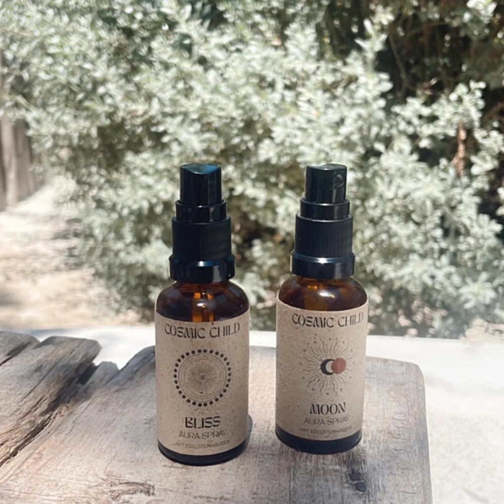 Braune Sprühflaschen von Cosmic Child Aura Spray Bliss und Moon stehen auf einer Holzplatte vor enem grünem Busch.