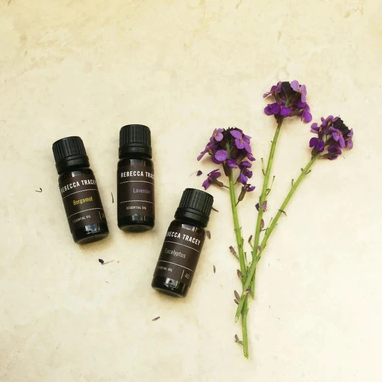 3 kleine dunkelbraune Glasflaschen Essential Oil von Rebeccy Tracey liegend, daneben Blumen.