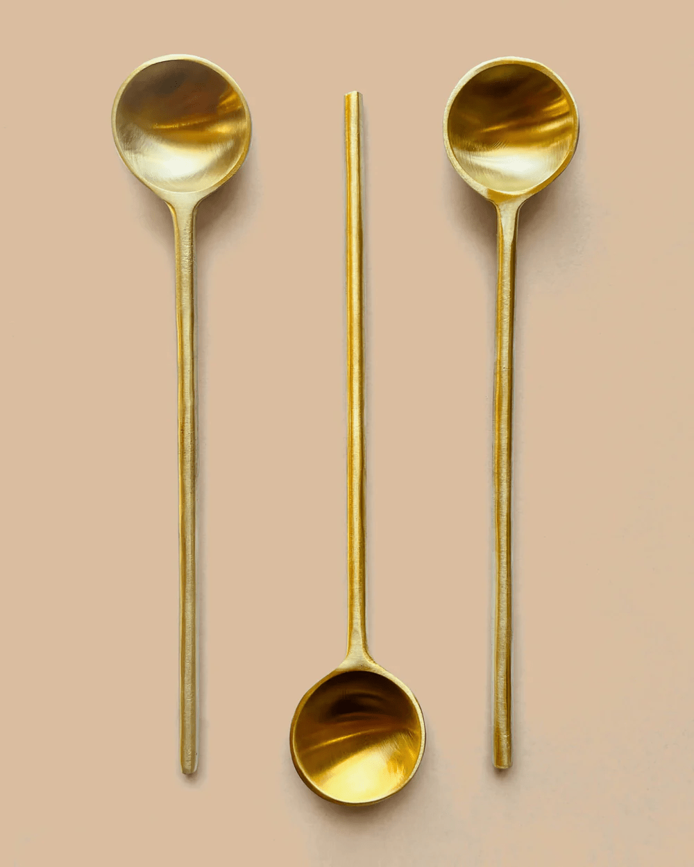 3 Messinglöffel - 2 mit dem Stiel nach unten, der Mittlere mit dem Stiel nach oben - liegen auf hellem Untergrund.