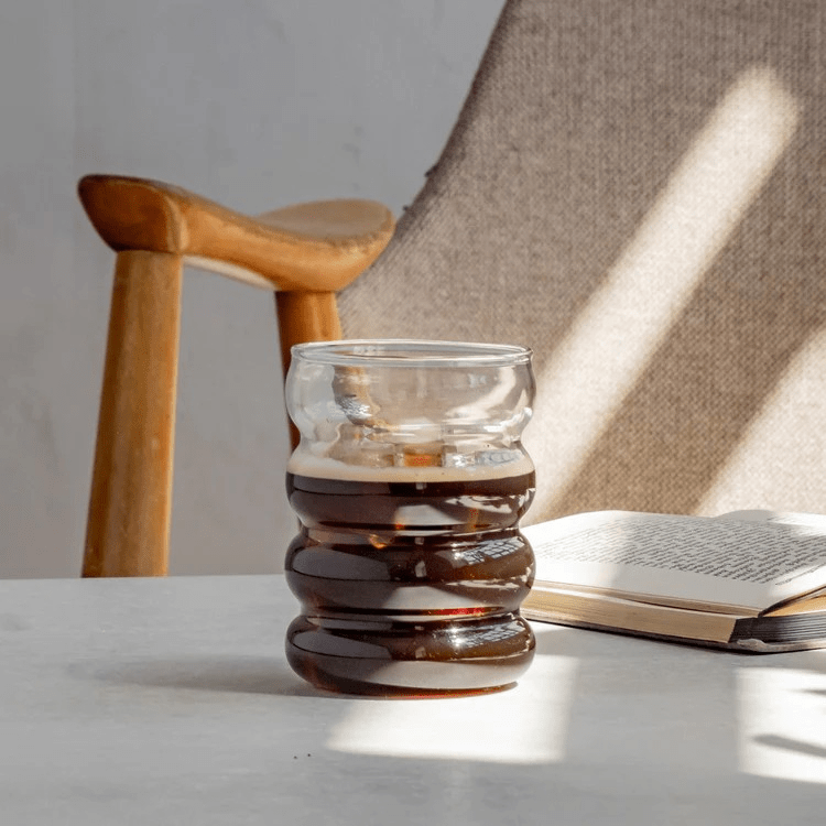 Mit Kaffee befülltes Bubble Glas Blanko von Eulenschnitt steht auf einem Tisch vor einem Stuhl und neben einem Buch. North Glow